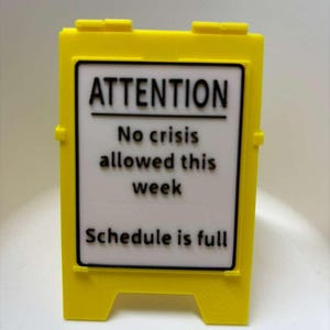 May include: A bright yellow sign with the words "ATTENTION" and the message "No crisis allowed this week. Schedule is full" in black text. The sign has a rectangular white background and a black border.