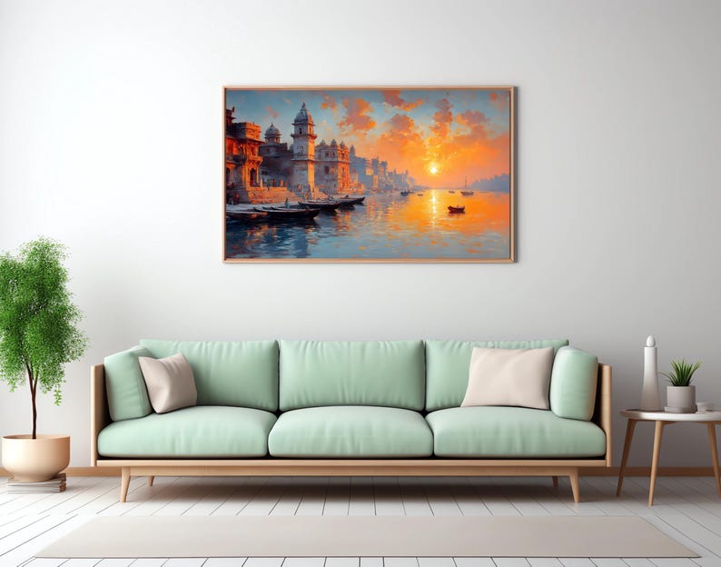May include: A framed landscape painting depicts a vibrant sunset over a river with buildings and boats. The artwork is displayed above a light green sofa with beige pillows, complemented by a small side table and a potted plant, creating a serene living room scene.