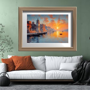 May include: A framed art print depicts a serene waterscape with buildings along the shore, boats, and a vibrant sunset. The artwork is set in a light brown wooden frame, displayed above a white sofa with orange pillows and a black throw blanket.