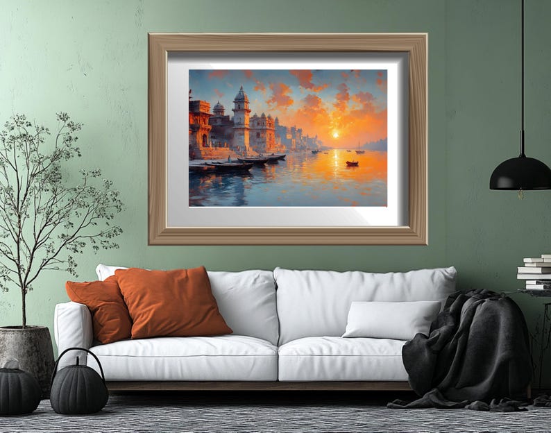 May include: A framed art print depicts a serene waterscape with buildings along the shore, boats, and a vibrant sunset. The artwork is set in a light brown wooden frame, displayed above a white sofa with orange pillows and a black throw blanket.