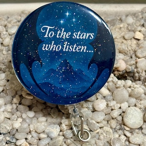 May include: A round, blue badge reel with the text "To the stars who listen..." in white script. The design features a starry night sky with mountains. The reel has a silver clip and a clear plastic strap.