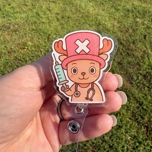 Puede incluir: Un portatarjetas transparente con un personaje de dibujos animados con un sombrero rosa y cuernos, sosteniendo una jeringa. El personaje tiene un estetoscopio alrededor del cuello. El portatarjetas tiene una correa de plástico transparente y un clip de metal.