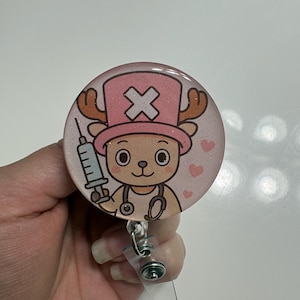 Puede incluir: Un carrete de insignia redondo y rosa brillante con un personaje de reno de dibujos animados que lleva un sombrero rosa con un símbolo de cruz. El personaje sostiene una jeringa y un estetoscopio. Pequeños corazones rosas están esparcidos alrededor del personaje.