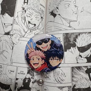 May include: A round button featuring three anime characters with vibrant colors and a glittery background. The button is set against a backdrop of manga panels in black and white.