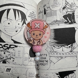 May include: A pink and white badge reel featuring a cartoon character with a pink hat and a syringe. The badge reel has a pink beaded accent and a silver clip. The background is a manga comic book page.