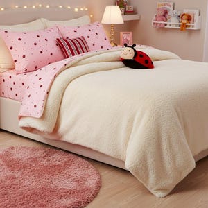 May include: A bedroom scene featuring a bed with pink ladybug-patterned bedding and a cream-colored comforter. A red and black ladybug plush toy rests on the bed. A pink rug and a white nightstand with a lamp complete the cozy setting.