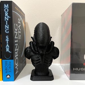 May include: A black Alien bust statue stands on a white surface, flanked by books. The statue has a detailed, textured design. The books include "Golden Son," "Morning Star," and "The Silo Series."