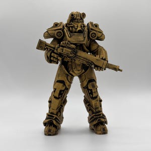 May include: A gold-colored figurine of a soldier in full tactical power armor, featuring a bulky helmet with circular eye lenses, heavy shoulder plating, and articulated leg armor. The figure holds a long rifle across its chest.