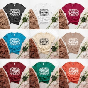 Peut inclure: Plusieurs t-shirts à manches courtes de différentes couleurs, dont noir, blanc, sarcelle, beige, bordeaux, marron, vert et orange. Chaque chemise affiche le texte "Your Design Here" dans une police stylisée. Les chemises sont présentées avec une couverture douillette, des fleurs et une tasse.