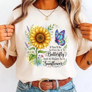 May include: A cream-colored t-shirt featuring a graphic print of a large yellow sunflower and a colorful butterfly. The design includes small wildflowers and the text: If You'll Be As Free As A Butterfly And As Happy As A Sunflower.