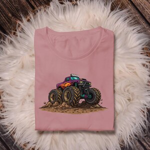 May include: A dusty rose-colored t-shirt featuring a cartoon illustration of a monster truck. The truck is purple with orange flames, large black tires, and green accents. The truck is kicking up dirt.