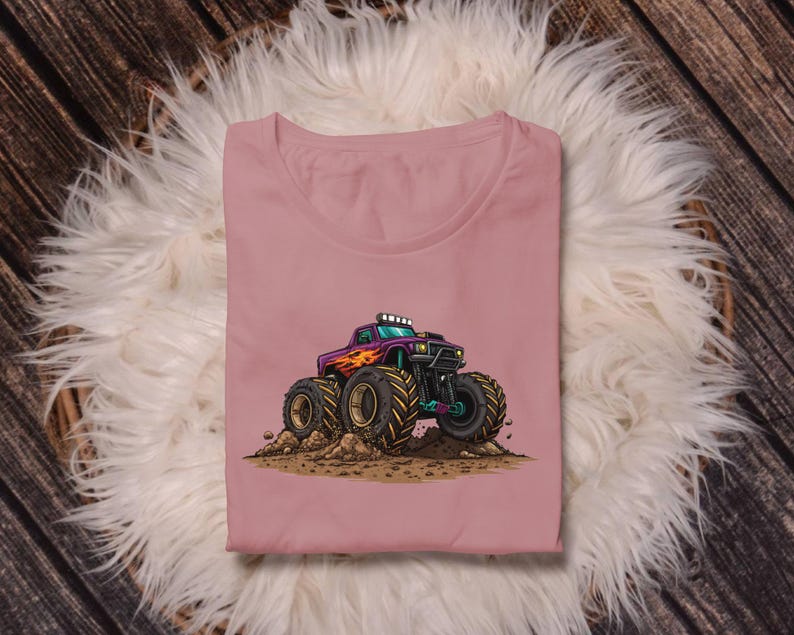 May include: A dusty rose-colored t-shirt featuring a cartoon illustration of a monster truck. The truck is purple with orange flames, large black tires, and green accents. The truck is kicking up dirt.