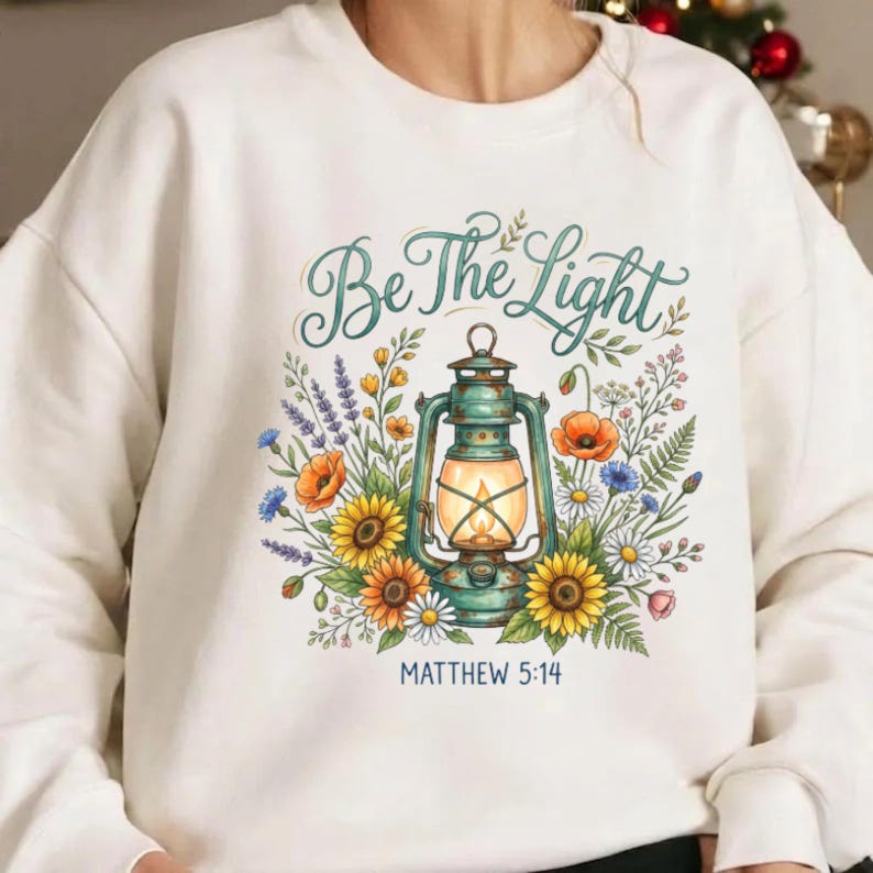 May include: A white crewneck sweatshirt featuring a graphic of a teal lantern surrounded by colorful wildflowers including sunflowers and daisies. The text "Be The Light" is printed in a teal script font above, with "MATTHEW 5:14" below.