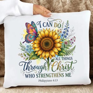 May include: A white square throw pillow featuring a large yellow sunflower, a colorful butterfly with orange, pink, and blue wings, and blue wildflowers. The text reads I CAN DO ALL THINGS THROUGH CHRIST WHO STRENGTHENS ME Philippians 4:13.
