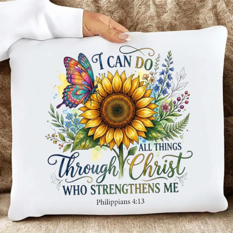 May include: A white square throw pillow featuring a large yellow sunflower, a colorful butterfly with orange, pink, and blue wings, and blue wildflowers. The text reads I CAN DO ALL THINGS THROUGH CHRIST WHO STRENGTHENS ME Philippians 4:13.
