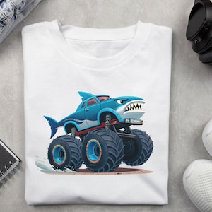 May include: White t-shirt featuring a cartoon monster truck designed as a shark. The truck is blue with a white and red accent, large black tires, and a shark fin. The shark truck has an angry expression.