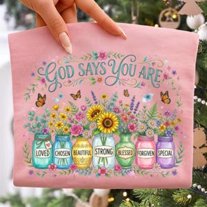 May include: A light pink t-shirt featuring a floral graphic with seven glass jars. The text reads "GOD SAYS YOU ARE" above the jars, which are labeled "LOVED," "CHOSEN," "BEAUTIFUL," "STRONG," "BLESSED," "FORGIVEN," and "SPECIAL" with butterflies.