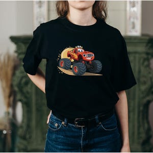 May include: Black t-shirt featuring a cartoon monster truck in red, orange, and yellow. The monster truck has large black tires and is smiling. The design is set against a yellow background.