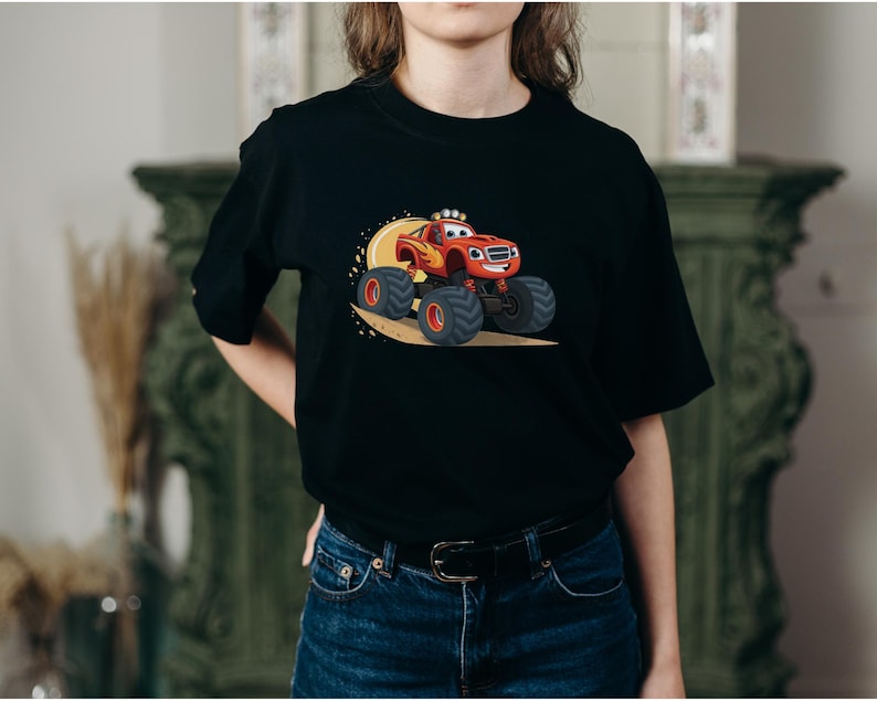 May include: Black t-shirt featuring a cartoon monster truck in red, orange, and yellow. The monster truck has large black tires and is smiling. The design is set against a yellow background.