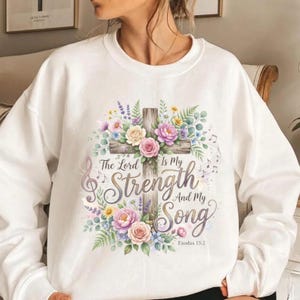 May include: A white crewneck sweatshirt featuring a central graphic of a wooden cross adorned with colorful flowers, including pink, yellow, and purple blossoms. The text reads, The Lord Is My Strength And My Song, with Exodus 15:2 below.