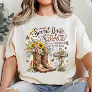 May include: A cream-colored t-shirt featuring a graphic of two brown cowboy boots filled with sunflowers, roses, and daisies. Next to the boots is a wooden cross adorned with flowers and butterflies. The text reads Saved By Grace and Redeemed by His Love.