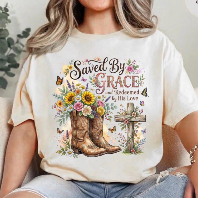 May include: A cream-colored t-shirt featuring a graphic of two brown cowboy boots filled with sunflowers, roses, and daisies. Next to the boots is a wooden cross adorned with flowers and butterflies. The text reads Saved By Grace and Redeemed by His Love.