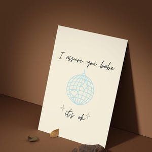 May include: A cream-colored print with the text "I assure you babe it's ok" in black script. A light blue disco ball graphic with stars and a heart is centered on the print. The print is leaning against a brown surface.