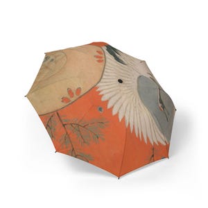 Puede incluir: Un paraguas abierto con un diseño de inspiración japonesa. El paraguas presenta una grúa con alas blancas sobre un fondo rojo y beige. El diseño incluye ramas y otros elementos naturales.