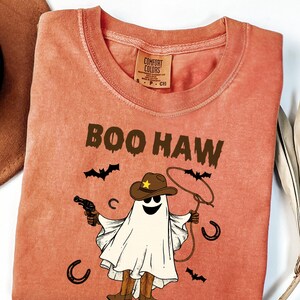 May include: Coral-coloured t-shirt featuring a ghost in a cowboy hat and boots, holding a gun and lasso. The text "BOO HAW" is above the ghost, with bats and horseshoes around the design.