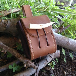 May include: A brown leather pouch with a flap and a bone toggle closure. The pouch is hanging from a brown leather strap and has a dark brown lining. The pouch is sitting on a pile of branches.