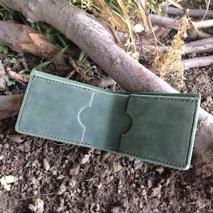 May include: A green leather wallet with a stitched edge and two slots for cards. The wallet is open and lying on the ground.