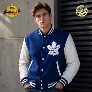 Op de afbeelding: Een marineblauw varsity-jack met witte mouwen en een witte geribbelde kraag, boorden en zoom met blauwe strepen. De voorkant toont een groot wit Toronto Maple Leafs-logo met de teamnaam in het blauw op de borst en witte knopen.