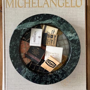 May include: A circular dark green marble ashtray with a mosaic interior base, containing several vintage matchbooks with various logos and text, resting on a large beige hardcover book titled MICHELANGELO in gold lettering.