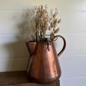 May include: A polished copper pitcher with a wide base, a curved handle, and a pointed spout. The pitcher is filled with a collection of dried beige grasses and fluffy seed heads, creating a natural arrangement within the metallic vessel.
