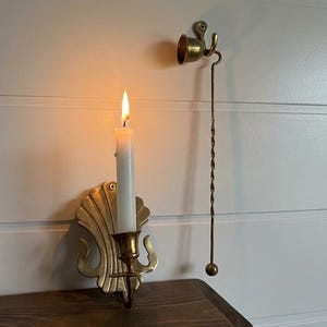 May include: A brass wall-mounted candle sconce with a shell-shaped backplate holding a lit white taper candle, accompanied by a matching brass candle snuffer featuring a long twisted handle and a small bell-shaped head mounted on a wall hook.