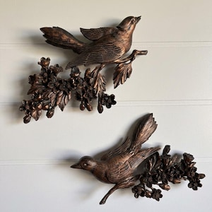 May include: Two wall-mounted decorative plaques featuring bronze-coloured birds perched on flowering branches. Each piece displays a bird with detailed feather textures and a cluster of small blossoms and leaves, finished in a metallic copper-bronze tone.