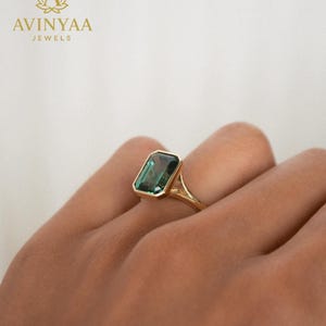 Może przedstawiać: Złoty pierścionek z prostokątnym, szmaragdowym, turkusowym kamieniem szlachetnym. Pierścionek ma prosty, elegancki design z cienką obrączką i oprawą w ramce. Widoczna jest nazwa marki "AVINYAA JEWELS".