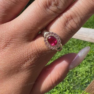 Puede incluir: Un anillo de oro con una piedra preciosa rosa vibrante de corte ovalado en el centro, rodeada por un halo de piedras rosas más pequeñas de corte redondo. El engaste tiene un diseño de borde festoneado y la banda es de oro pulido brillante.