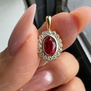May include: A gold pendant featuring a central oval-cut red ruby surrounded by a halo of small round white diamonds. The pendant has a scalloped edge setting and a gold bail at the top. The text Avinyaa jewels is visible across the image.