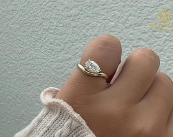 May include: A gold ring featuring a pear-cut diamond. The diamond is set in a swirling design. The ring is on a finger, against a light green wall.
