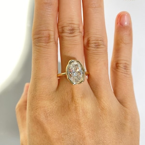 May include: A gold ring with an oval-shaped, clear gemstone. The gemstone is set in a bezel setting with a unique, irregular shape. The ring is displayed on a hand, showcasing the jewelry's design and sparkle. The brand name "AVINYAA JEWELS" is visible.