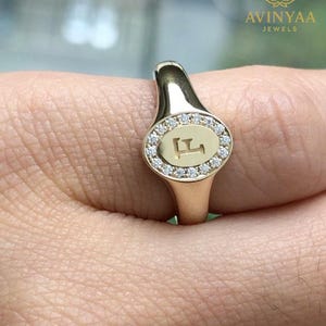 Pode incluir: Um anel de sinete dourado com uma face oval cravejada de diamantes. A face apresenta a letra "F" no centro. O anel é usado no dedo. A marca "AVINYAA JEWELS" é visível no fundo.