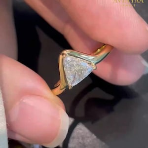 Könnte beinhalten: Ein goldener Ring mit einem klaren, dreieckigen Edelstein. Der Ring hat ein einzigartiges, geschwungenes Design. Der Markenname "AVINYAA JEWELS" ist oben rechts sichtbar.