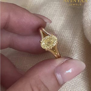 May include: A gold ring with a large, oval-cut yellow gemstone. The ring's band has a split design, with the text "AVINYAA JEWELS" visible. The ring is held up by a person's fingers.