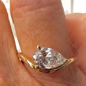 Könnte beinhalten: Ein goldener Ring mit einem birnenförmigen Diamanten. Der Diamant ist in einem geschwungenen Band gefasst. Der Ring wird an einem Finger gezeigt. Der Markenname "AVIN YAA JEWLS" ist sichtbar.