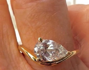 以下が含まれることがあります： 洋梨型のダイヤモンドをあしらったゴールドリング。 ダイヤモンドは、曲線的なバンドにセットされています。 リングは指に装着されています。 ブランド名「AVIN YAA JEWLS」が表示されています。