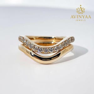 Puede incluir: Dos anillos dorados apilados. El anillo superior tiene un diseño curvo y está incrustado con pequeñas piedras preciosas transparentes. El anillo inferior es una banda lisa y curva. El fondo es blanco. El texto "AVINYAA JEWELS" está en la esquina superior derecha.