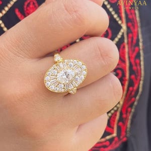 Puede incluir: Un anillo dorado con un diseño ovalado. El anillo está adornado con piedras claras que rodean una piedra central, creando un efecto halo. Una joya con una banda dorada.