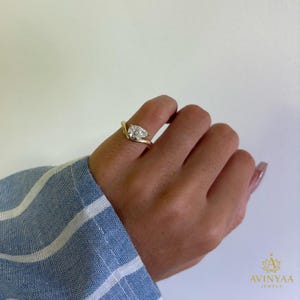 May include: A gold ring featuring a pear-shaped diamond cluster. The ring's band has a curved design. The ring is displayed on a hand, with a blue and white striped shirt visible in the background. The ring is a piece of jewellery.