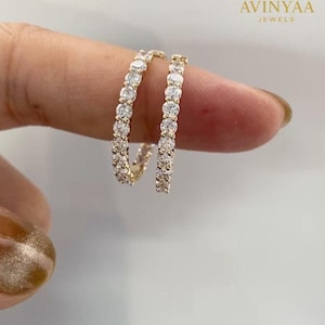 May include: A pair of gold hoop earrings, each encrusted with a row of sparkling, clear gemstones. The earrings are held up by a finger, showcasing the jewelry's design. The logo "AVINYAA JEWELS" is visible in the upper right corner.